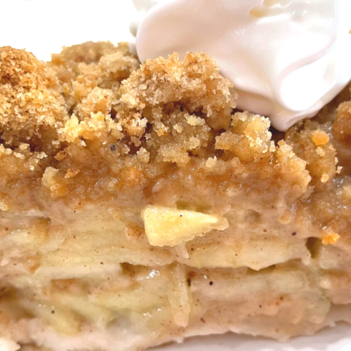 CLOSE UP SIDE PICTURE OF A SLICE OF DUTCH APPLE PIE WITH A DOLLOP OF COOL WHIP ON TOP AS IT SITS ON A WHITE PLATE.