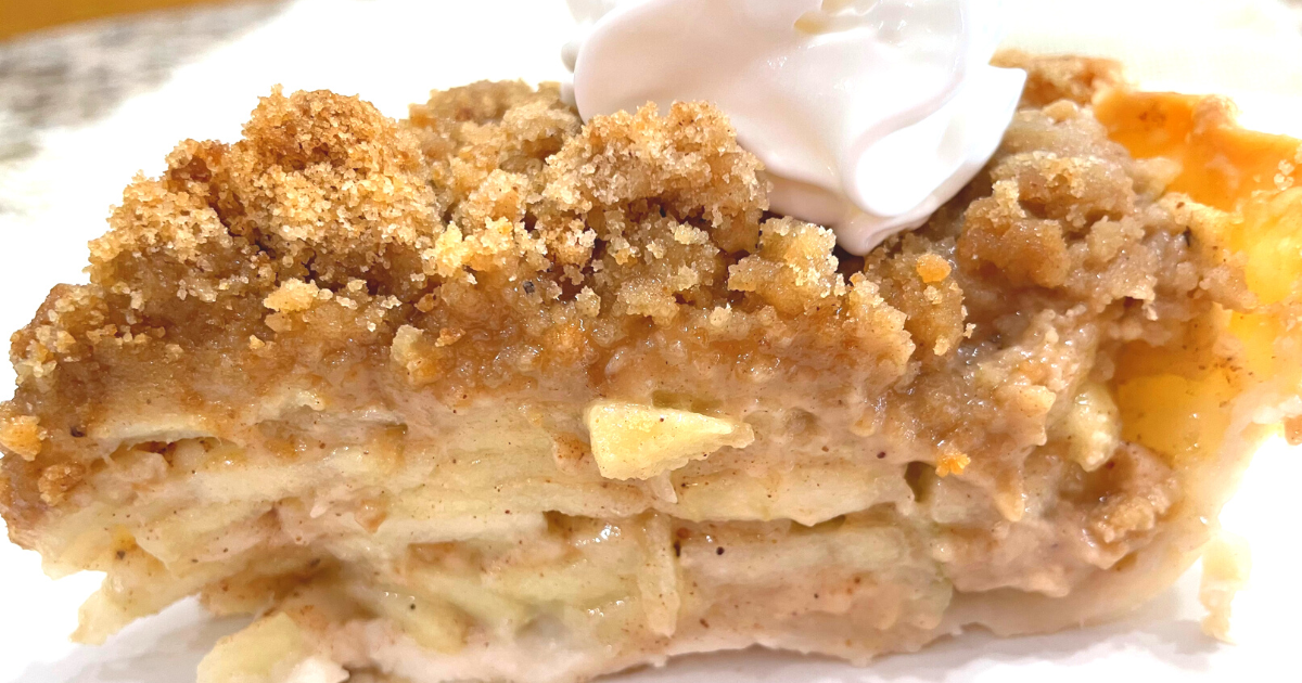CLOSE UP SIDE PICTURE OF A SLICE OF DUTCH APPLE PIE WITH A DOLLOP OF COOL WHIP ON TOP AS IT SITS ON A WHITE PLATE.