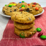 A stack of monster cookies on a red, cloth napkin in front of a white plate full of monster cookies
