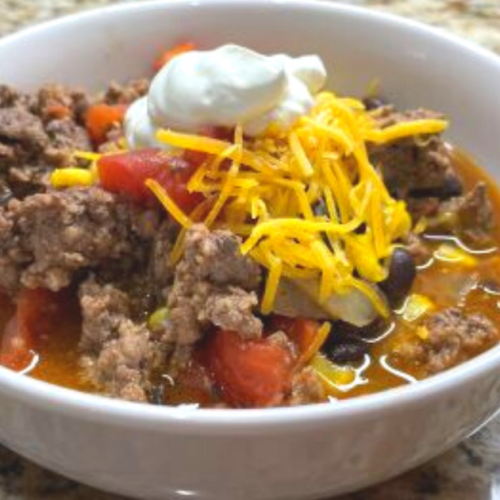 White bowl full of taco soup topped with shredded cheese and a dollop of sour cream.
