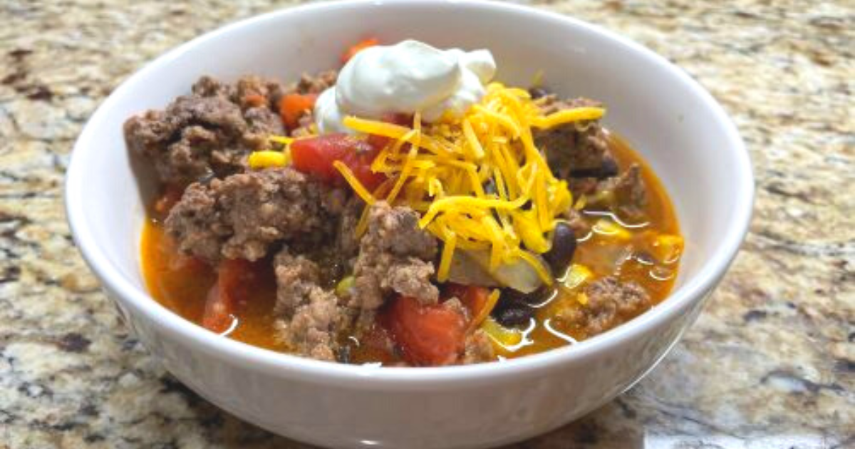 White bowl full of taco soup topped with shredded cheese and a dollop of sour cream.