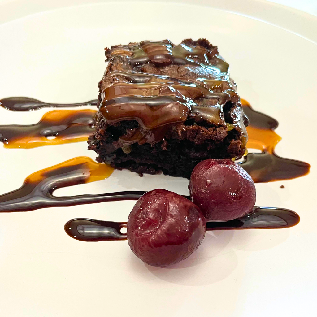 Brownie on a white plate drizzled with caramel and chocolate and two cherries