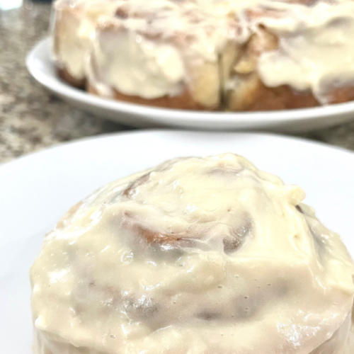 Homemade cinnamon roll smothered in icing on a white plate with the rest of the cinnamon rolls in the background.