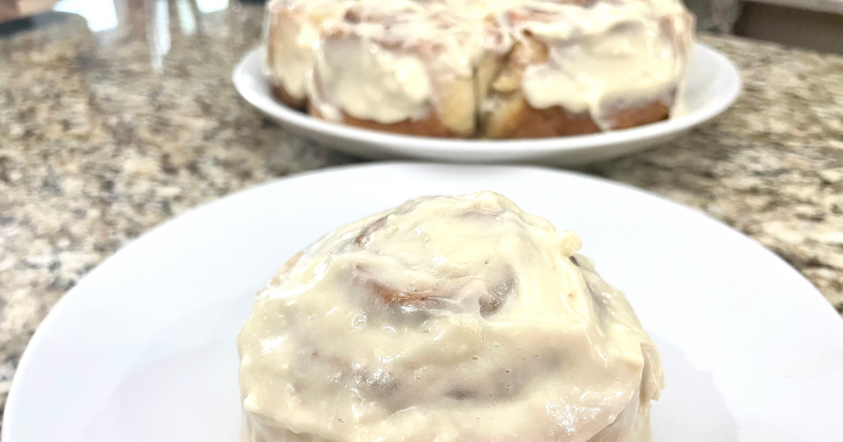 Homemade cinnamon roll smothered in icing on a white plate with the rest of the cinnamon rolls in the background.