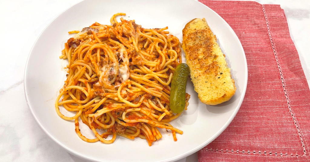 Image of a serving of the fried spaghetti on a white plate with a garlic bread stick and a pickle.
