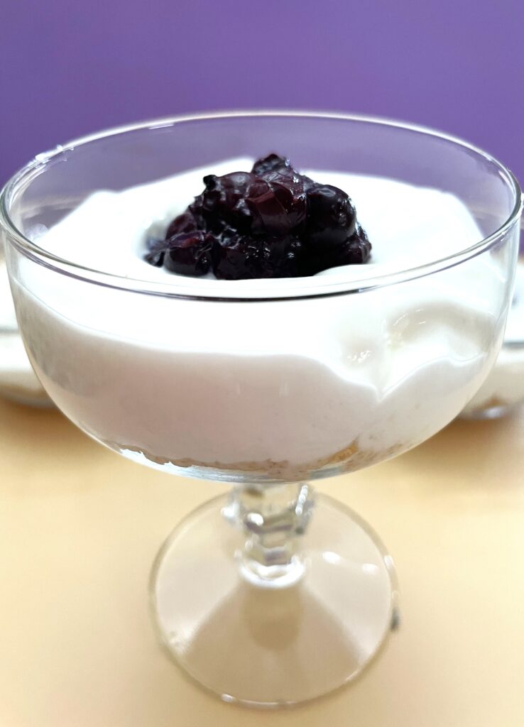 Close up of a stem glass with the graham cracker crumbs on the bottom, cream cheese dessert in the middle topped with blueberries in the center.