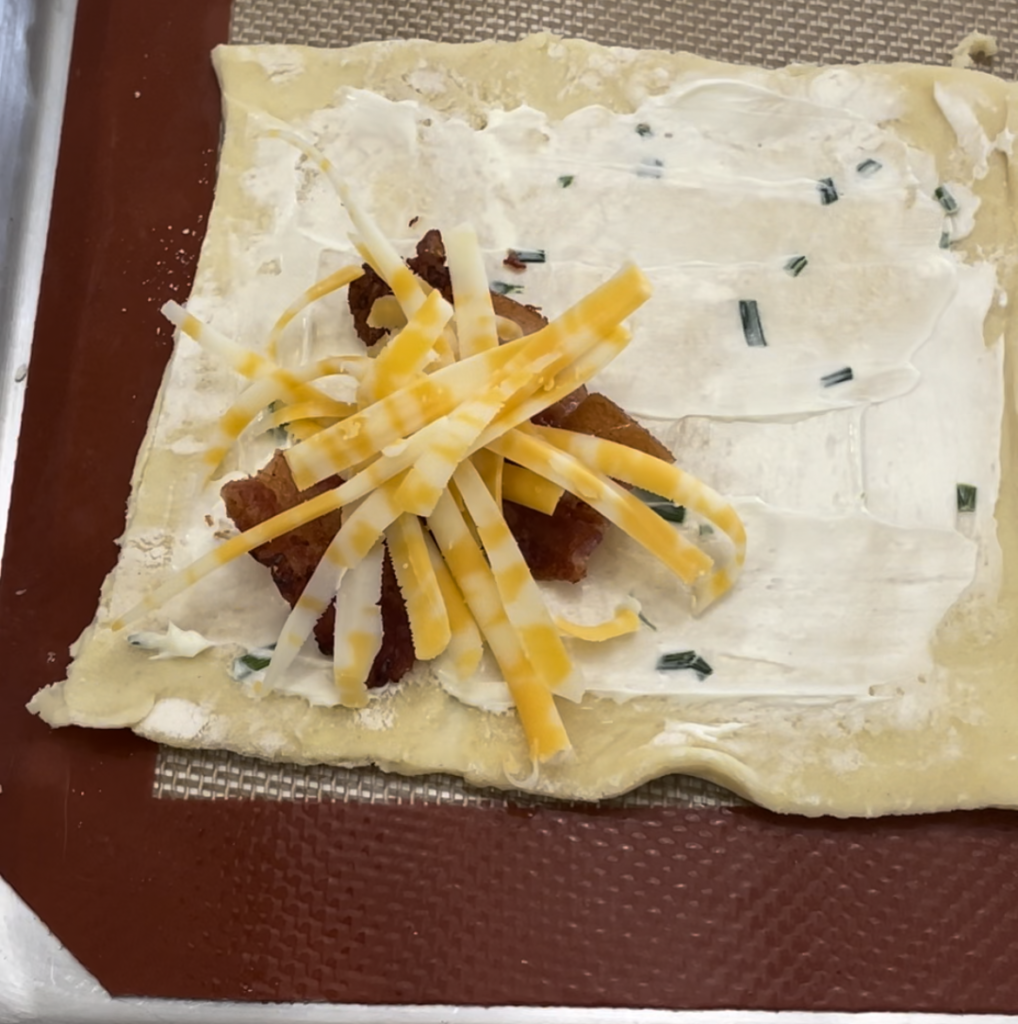 Puff pastry square with cream cheese and chives spread on it and a couple pieces of bacon and shredded cheese in one corner before closing it up.