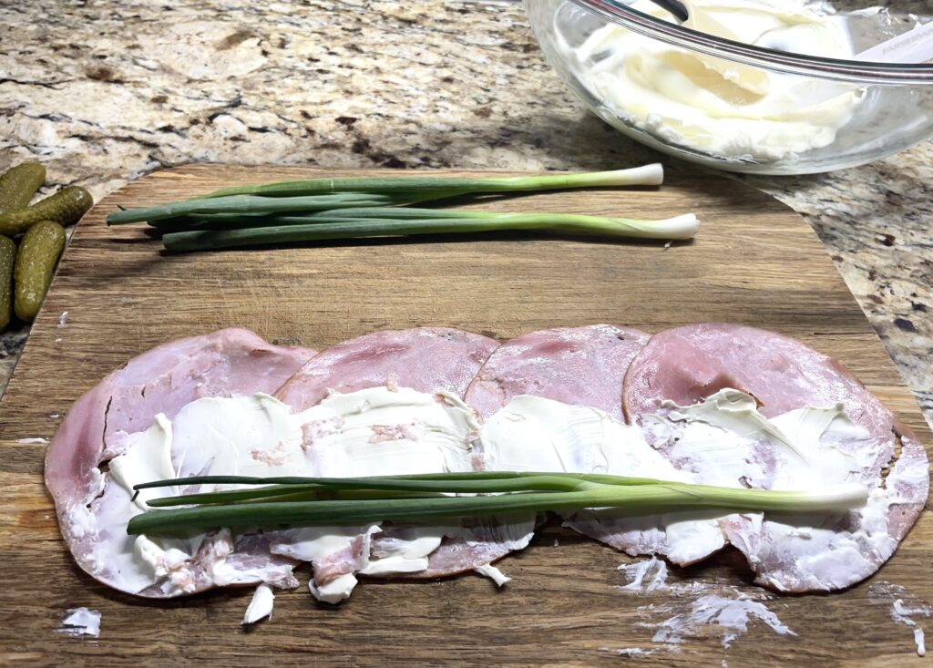 Ham slices layered on a wooden cutting board. Cream cheese spread out over them and an onion along the side ready to be rolled up.