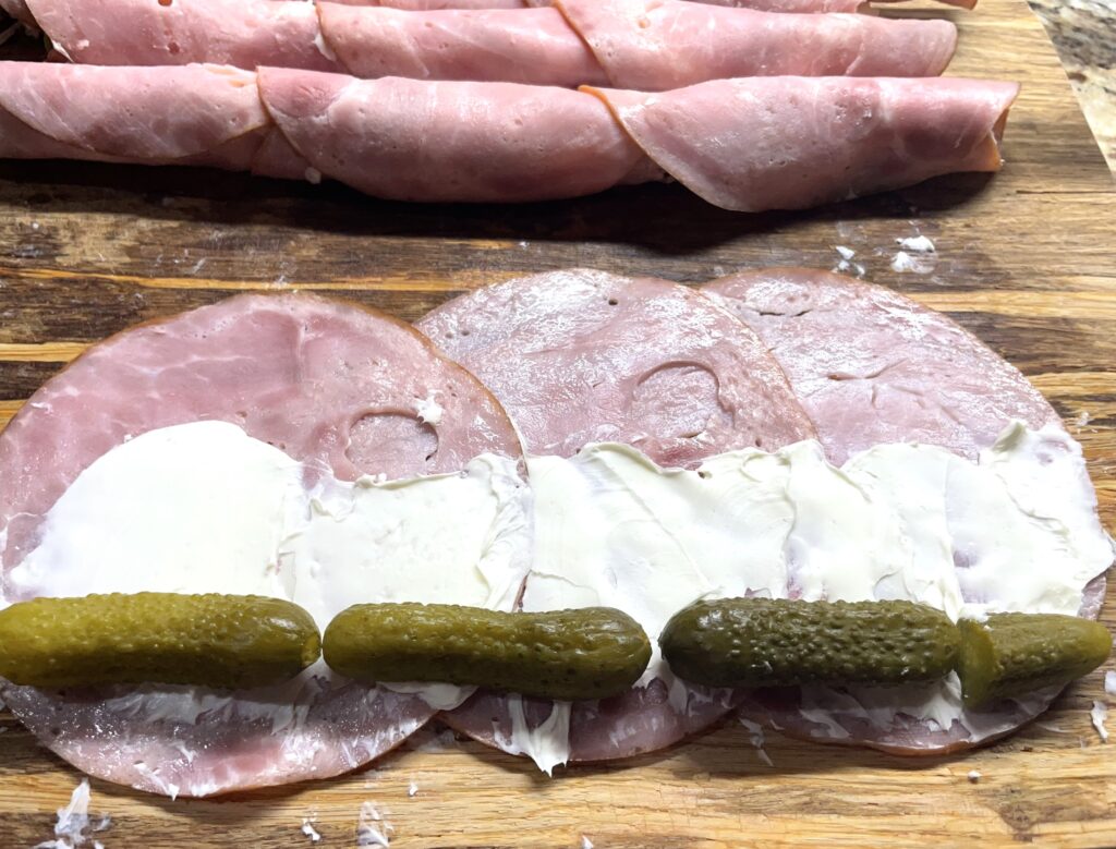 two roll up logs completed and three slices of ham layered with cream cheese spread on them with pickles lined up along one side ready to roll up.
