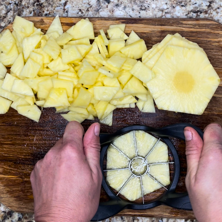 Using an apple corer slicer on a wooden cutting board with a pile of already cut up pineapple and whole pineapple rounds in the picture.