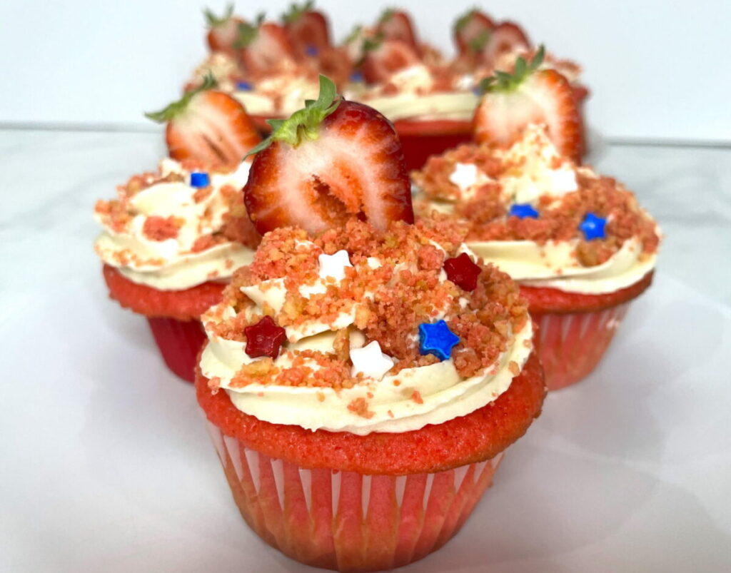 Several Strawberry Crunch Cupcakes completed with icing and crunch. Then they are topped with a strawberry half and little red, white, and blue candy stars.